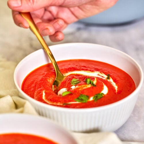 Roasted Red Pepper Soup 2 Hand holding a spoon scooping creamy roasted red pepper soup from a bowl, showing rich texture and garnished with herbs.