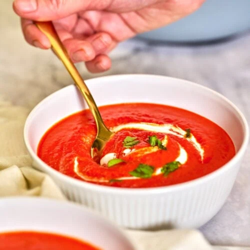 Roasted Red Pepper Soup 2 Hand holding a spoon scooping creamy roasted red pepper soup from a bowl, showing rich texture and garnished with herbs.
