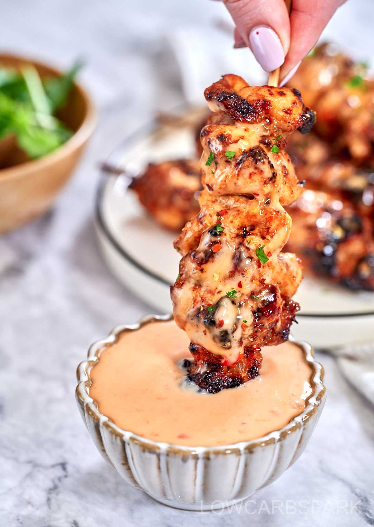 A hand with pink nails holds a chicken skewer over a small bowl of pink sauce. The cooked chicken has bits of green herbs and some charred spots. More skewers and a green salad are out of focus in the background. 