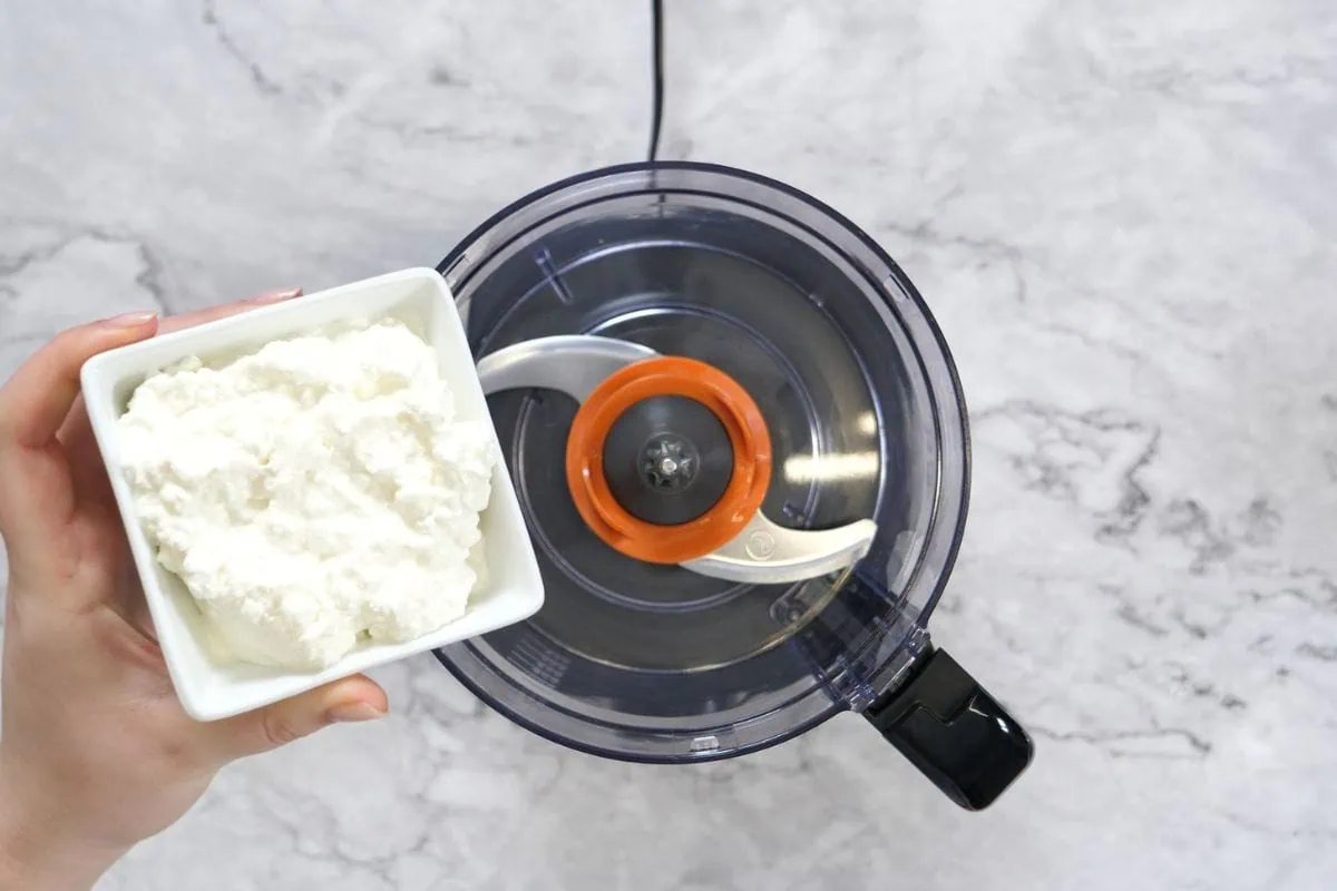Cottage Cheese Protein Muffins 5 A bowl of cottage cheese being held over a food processor, ready to be blended into muffin batter.