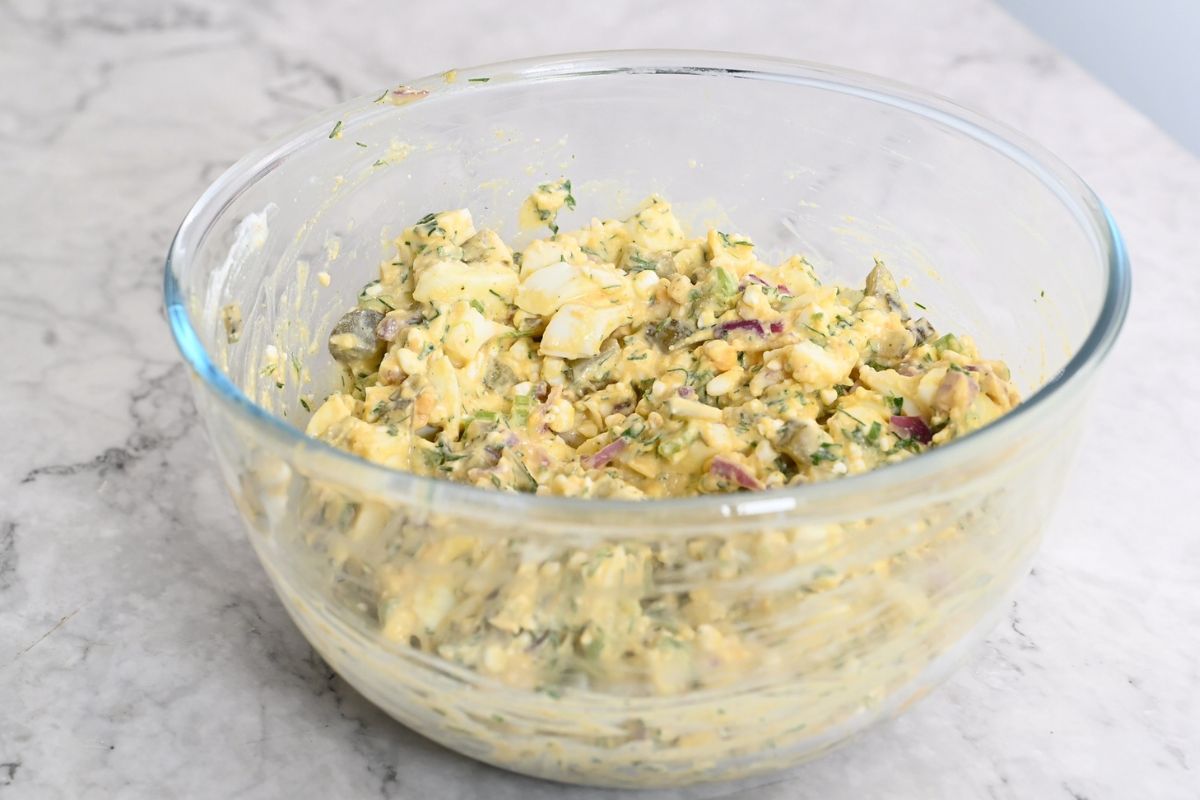 Creamy chopped-egg salad mixed with pickles, celery and dill in clear mixing bowl.
