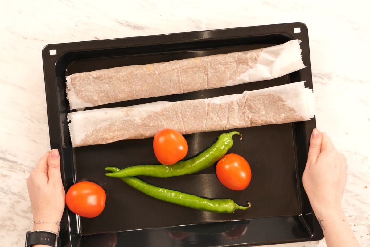 Two parchment-wrapped kebab meat logs placed on a baking tray with tomatoes and green peppers.