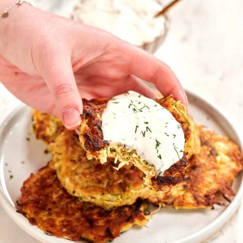 Cabbage Fritters 9 Hand holding a crispy golden cabbage fritter topped with creamy dill sauce over a plate of low carb cabbage fritters.