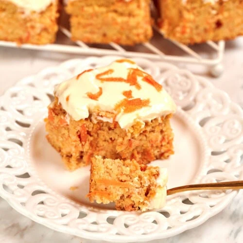 Cottage Cheese Carrot Cake Bars 12 High-protein cottage cheese carrot cake bars topped with creamy frosting and shredded carrots on a white plate.