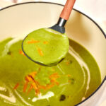 Ladle lifting creamy broccoli cheddar soup from a pot garnished with shredded cheddar cheese.