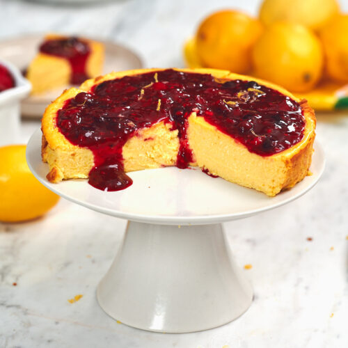 Cottage Cheese Lemon Cheesecake 14 Whole cottage cheese lemon cheesecake topped with glossy mixed berry sauce and fresh lemon zest on a white cake stand.