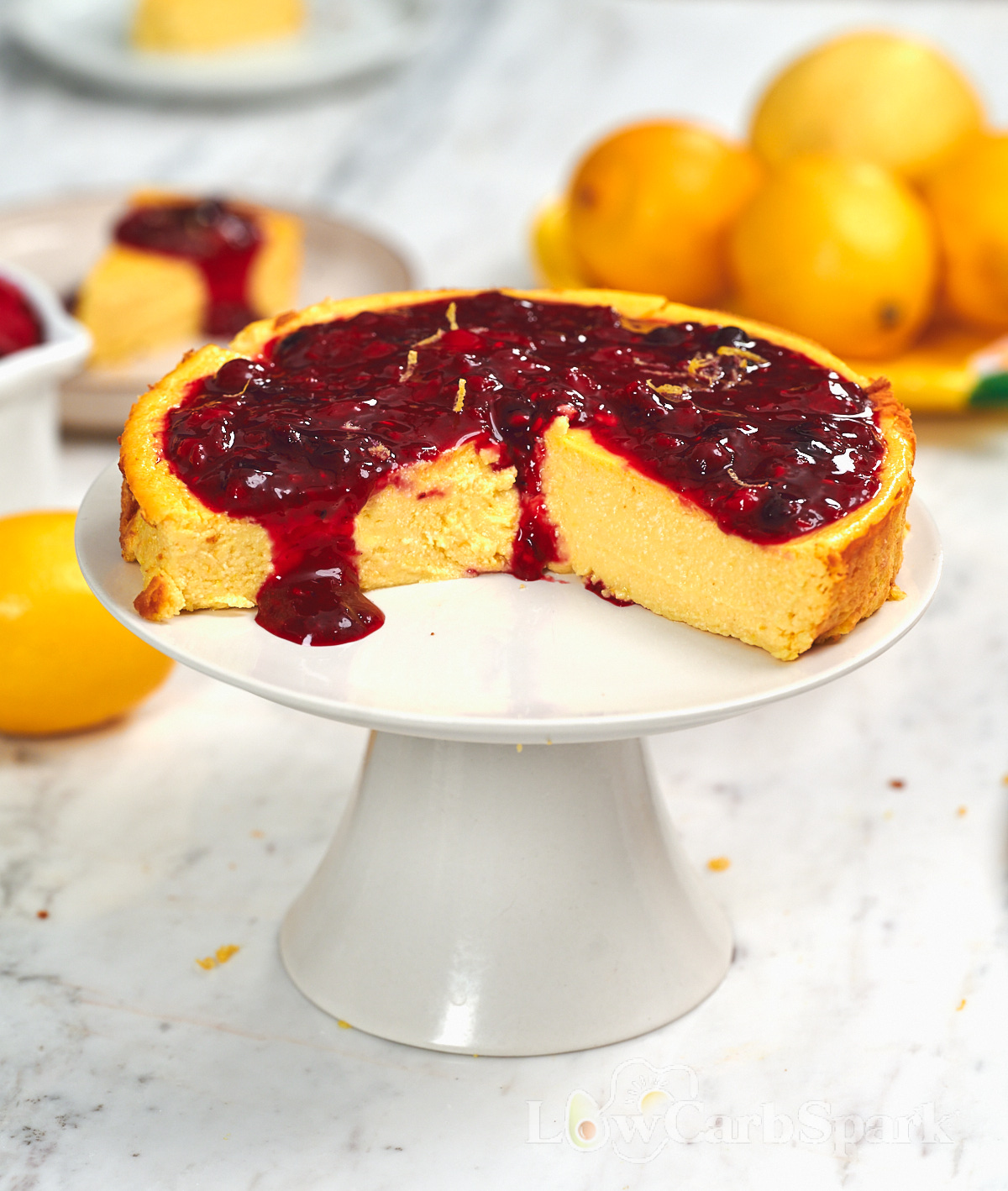 Cottage Cheese Lemon Cheesecake 1 Whole cottage cheese lemon cheesecake topped with glossy mixed berry sauce and fresh lemon zest on a white cake stand.