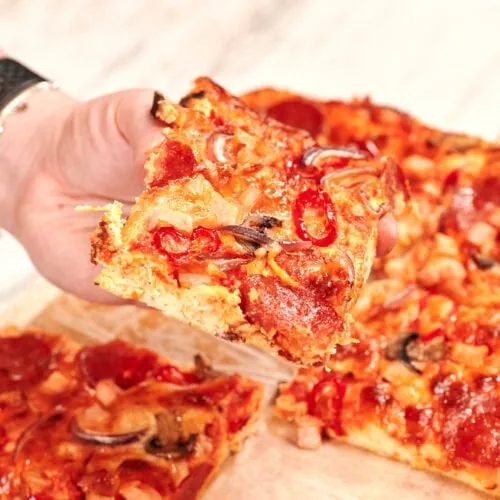 Sheet Pan Cottage Cheese Pizza 9 Hand holding a slice of sheet pan cottage cheese pizza with pepperoni, mushrooms, red onion, and melted cheese on a thick high-protein crust.