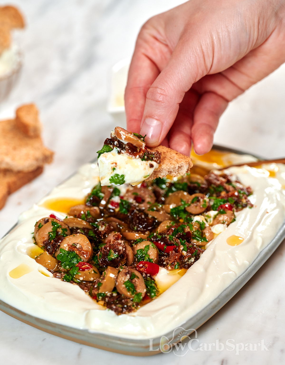 Whipped Cottage Cheese Dip with Sun-Dried Tomatoes 2 Hand dipping a slice of toasted bread into creamy whipped cottage cheese dip with a flavorful topping of sun-dried tomatoes, olives, garlic, parsley, and olive oil.