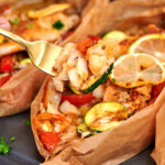 Fork holding a bite of Mediterranean baked fish with zucchini and roasted vegetables from a parchment paper boat.