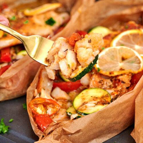 Mediterranean Baked Fish Boats 13 Fork holding a bite of Mediterranean baked fish with zucchini and roasted vegetables from a parchment paper boat.