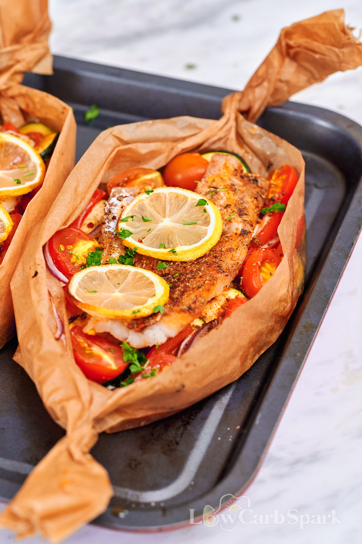 Mediterranean Baked Fish Boats 12 Mediterranean baked fish in a parchment boat with roasted vegetables, lemon slices, and fresh herbs on a baking sheet.