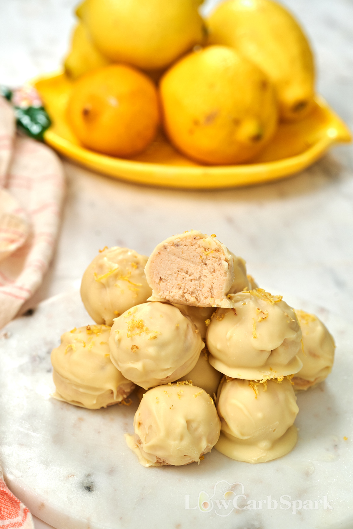 Lemon Cottage Cheese Truffles 11 Stack of homemade lemon cottage cheese truffles with white chocolate coating and lemon zest, with fresh lemons in the background.